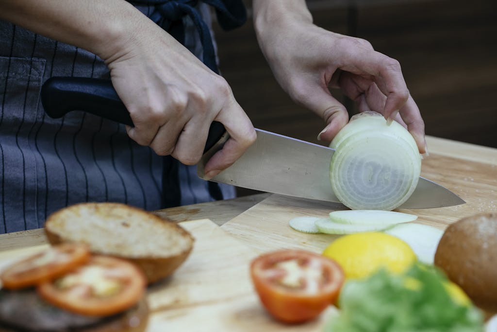 Unrecognizable female cook slicing onion on cutting board at table with blurred tomatoes and toasted buns in kitchen during cooking process